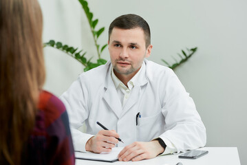 A portrait of a doctor in a white lab coat who is sitting at a desk and listening to a female...