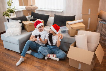 Newlywed couple celebrate Christmas or New Year in their new apartment. Young happy man and woman drinking wine, celebrating moving to new home and sitting among boxes.