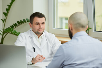 A caucasian doctor in a white lab coat is sitting at a desk and listening to a story of a bald male...