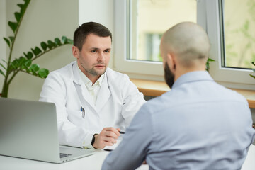 Naklejka na ściany i meble A caucasian doctor in a white lab coat is sitting at a desk and carefully listening to a story of a bald male patient in a hospital. A man with a beard at an appointment in a doctor's office.