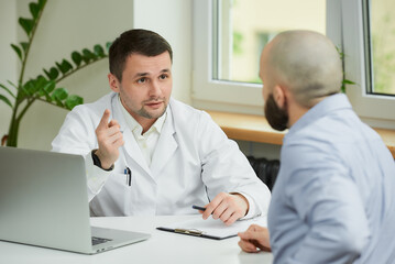 A caucasian doctor in a white lab coat is sitting at a desk and describing the treatment to a bald male patient in a hospital. A man with a beard at an appointment in a doctor's office.