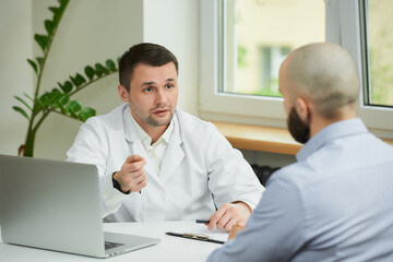 A caucasian doctor in a white lab coat is describing and demonstrating the treatment to a bald male patient in a hospital. A man with a beard at an appointment in a doctor's office.