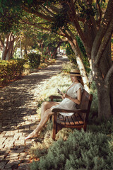 A girl with a book in her hands sits on a bench in the shade of picturesque trees. Lifestyle. Young woman resting from the summer heat in the shade of trees, resort village Gocek, Turkey