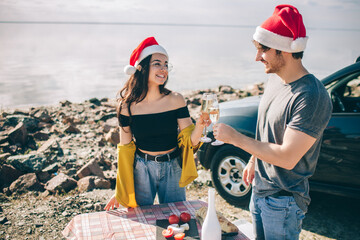 Travel, tourism - Man and woman Celebrates New Year or Christmas and drink champagne near the water at a folding portable table. Picnic near the water. Couple going on Adventure. Car travel concept.