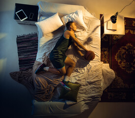 Catching. Top view of young professional basketball player sleeping at his bedroom in sportwear with ball. Loving his sport even more than comfort, playing match even if resting. Action, motion, humor