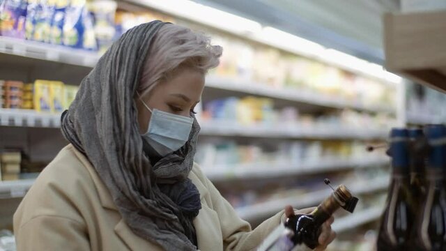 Young woman in a medical protective mask chooses wine at a grocery store. Shopping, quarantine, coronavirus. Health customer care.