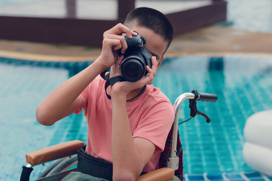 Camera in hands of Asian special child on wheelchair take photo at swimming pool in front of sea beach,Boy happiness in holidays with family time on the travel,Lifestyle of happy disabled kid concept.