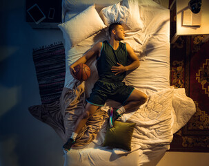 Fototapeta na wymiar Champion. Top view of young professional basketball player sleeping at his bedroom in sportwear with ball. Loving his sport even more than comfort, playing match even if resting. Action, motion, humor