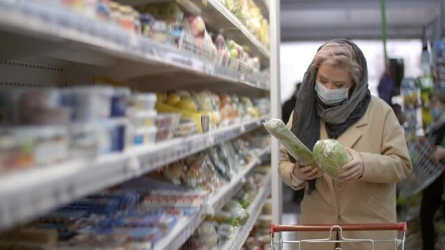Young woman wearing a medical protective mask picks vegetables at a grocery store during a pandemic, then puts the goods in a shopping cart. Shopping, quarantine, coronavirus. Health customer care.