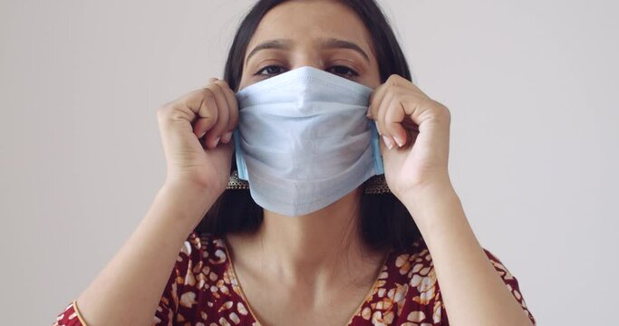 Young Indian woman wearing a medical surgical mask in covid pandemic times of new normal following rules and regulations - ill flu cough unwell adjusting mask worried concerned symptoms aware spread