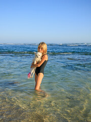 Cute little girl holding small white chihuahua dog with pink tail on the beach. Childhood concept. Spending time outdoor. Ocean during low tide. Bali, Indonesia