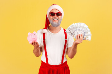 Fototapeta na wymiar Positive adult grey bearded man in santa claus hat and trendy sunglasses holding dollar banknotes and piggy bank, savings, high rate deposit. Indoor studio shot isolated on yellow background