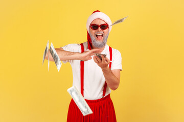 Positive generous grey bearded man in santa claus hat, sunglasses and pants with suspenders throwing dollar cash into air, celebrating winter holidays. Indoor studio shot isolated on yellow background - Powered by Adobe