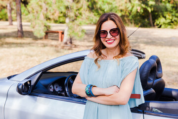 Attractive woman leaning and posing at convertible car. Happy pretty girl. Outdoor fashion...