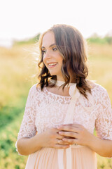 Fototapeta na wymiar portrait of pregnant european brunette woman girl in pastel pink dress with hat on the field during sunset