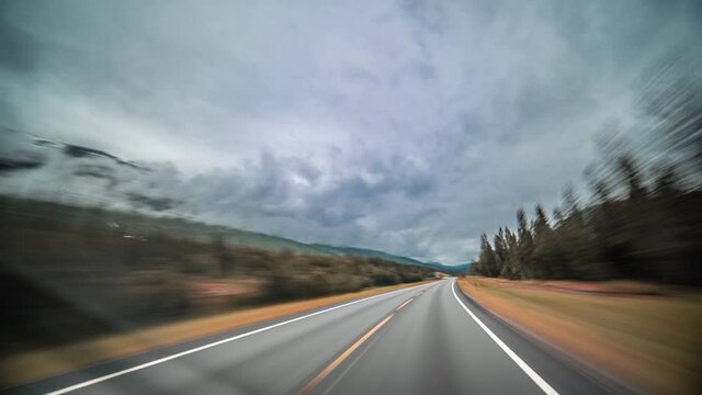 A drive on a E39 somewhere in Northern Norway. Narrow two lane freeway leading through the tundra. Moss, lichen and grass colored in orange and brown cover the landscape. Gray clouds covering the sky.