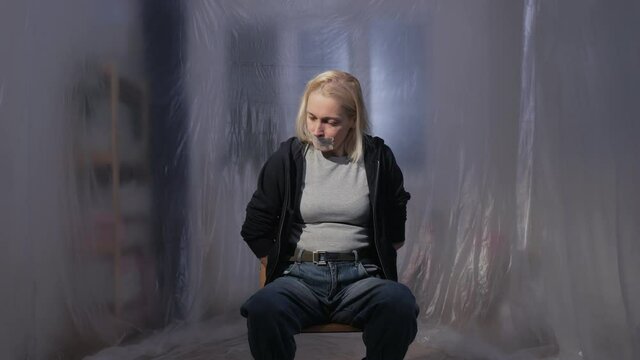 Female hostage sitting on a chair in a room with her mouth taped shut