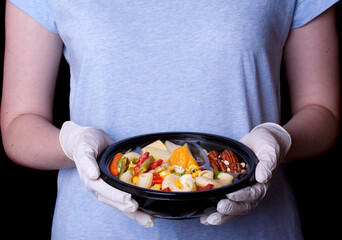 Pasta salad, slices of fruit and mix of nuts in the disposable bowl held by hands in gloves
