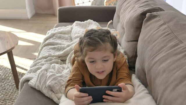 A little girl with a beautiful hairstyle attentively watches cartoons online on her mobile phone lying on the sofa and covered with a blanket