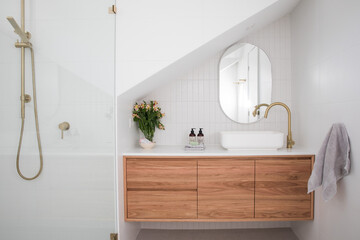 Brushed brass tap mixer on timber vanity with white basin bowl against white tiled wall in a new modern elegant bathroom lit by natural light from a nearby window modern interior house renovation new  - obrazy, fototapety, plakaty