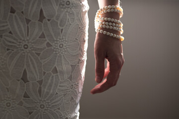 A woman wearing a string of pearls as a bracelet a pearl covered purse dressed in a white lace dress lit by natural light from a nearby window casting shadows