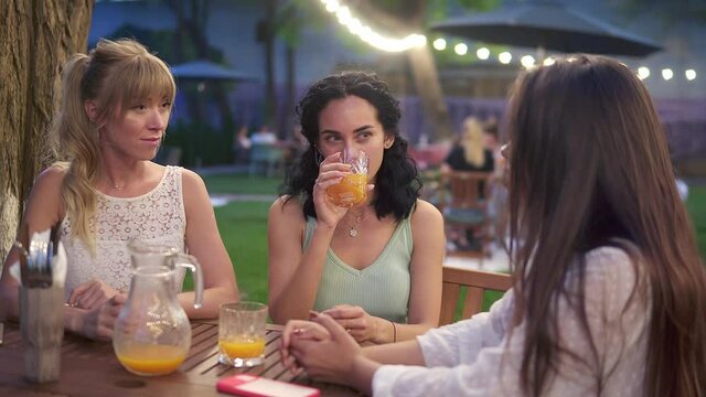 Three elegant female friends sit in the outdoors cafe, drink juice and have fun communicating