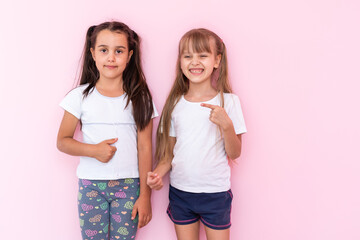 Amazed little kids girls showing white t-shirt, isolated on pink background. Childhood lifestyle concept. Mock up copy space. Pointing index finger aside