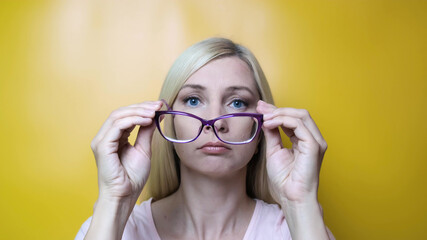 A middle age blond woman putting on stylish fashionable glasses and smiling to camera against yellow background, myopia, astigmatism and ophthalmologic diseases comcept