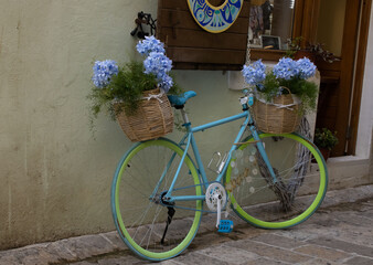 Fototapeta na wymiar Bright blue bike with light green wheels. Two baskets with flowers. Bicycle at the wall of an old house