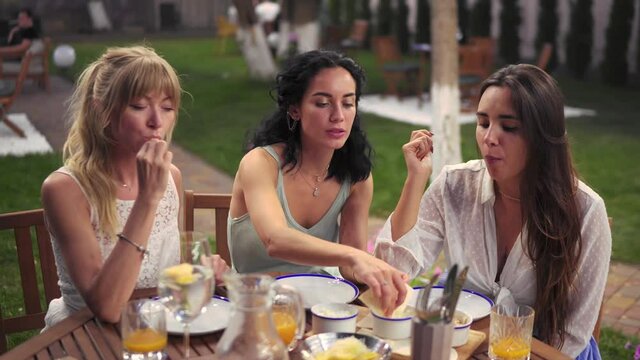 Three cheerful women resting talking drinking, eating in summer veranda cafe casual city friendship models outdoors