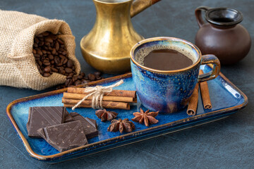 A cup of coffee, cinnamon sticks, chocolate, star anise, coffee beans, a coffee maker and a creamer in a beautiful dish against a dark table background. Still life.