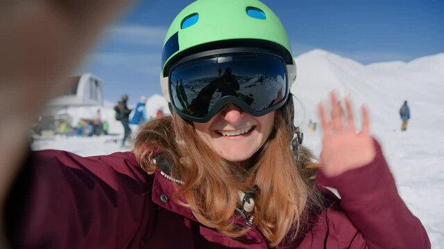 Happy young woman in green rider helmet and big sun goggles waves to camera on video call or live stream on social media. Ski resort winter vacation hotel, view on ski slopes, female snowboarder smile