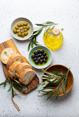 Green and black olives with olive oil in a glass bottle, olive tree sprigs and cut fresh ciabatta bread on wooden cutting board. White rustic background, mediterranean food concept, top view