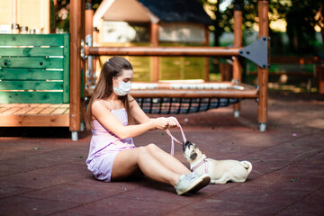 Happy dog. A girl in a mask holds a dog in her arms. Young woman walking with a pug dog in summer park. Portrait of a pug. Portrait of a beautiful pug puppy dog. The dog is lying on the ground.