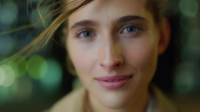 Closeup portrait of naturally beautiful young woman looking at camera and smiling surrounded by bokeh at night, her hair waving in wind in slow motion
