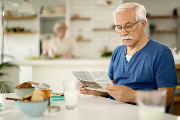 Mature man reading newspaper while having breakfast at home.