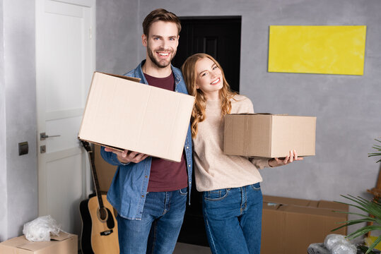 joyful man and woman holding carton boxes in new home