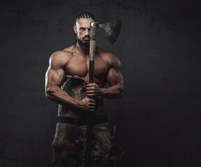 Fototapeta na wymiar Bodybuilder in fashion of furious viking posing holding axe near face with naked torso in dark background.