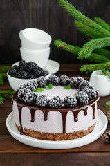 Blackberry cream mousse cake (no baked cheesecake) with chocolate glaze and frozen blackberry on top on a white plate on a wooden background. Vertical, copy space.