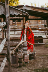 A beautiful Slavic girl with long blonde hair and brown eyes in a white and red embroidered suit is sitting next to a wooden house.Traditional clothing of the Ukrainian region.