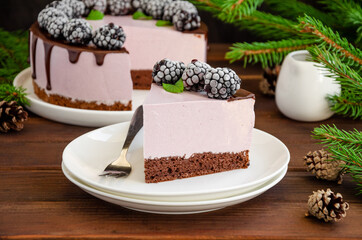 Blackberry cream mousse cake (no baked cheesecake) with chocolate glaze and frozen blackberry on top on a white plate on a wooden background. Horizontal, copy space.