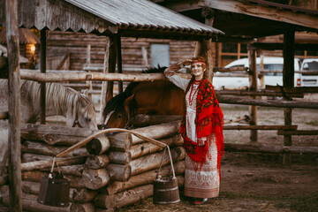 A beautiful Slavic girl with long blonde hair and brown eyes in a white and red embroidered suit is sitting next to a wooden house.Traditional clothing of the Ukrainian region.