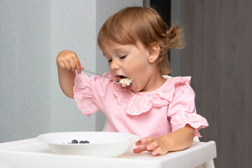 Little kid girl eating her morning porridge oatmeal with blueberries herself, sitting in baby chair in kitchen