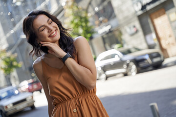 Perfect summer day. Portrait of a young attractive happy woman wearing dress keeping eyes closed and smiling while walking on the city streets on a sunny day