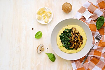 A portion of polenta or corn grits with fried mushrooms and stewed spinach in a gray plate on a light concrete background. Polenta or corn grits recipes.