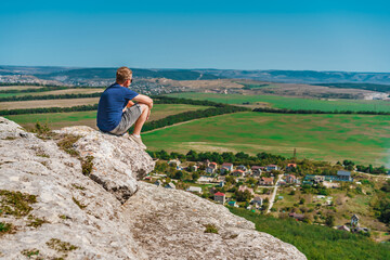 Naklejka na ściany i meble A blond man sits on a high cliff, enjoying the view of the green valley