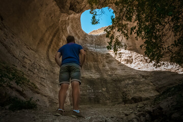 Rear view of a young man adventurer standing in an underground cave with an hole to the outside, sunlight through a cave