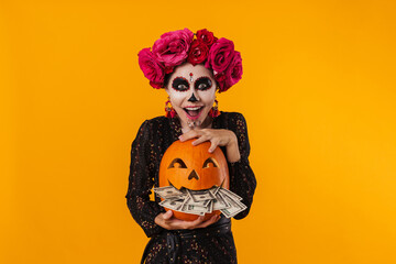 Delighted girl in halloween makeup posing with pumpkin and dollars