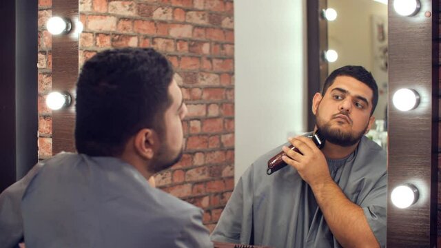 Caucasian man with machine and comb trimming himself beard front mirror. Bearded man grooming beard by electric trimmer in barber shop