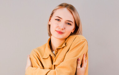 Smiling pretty young woman hugging herself and looking at camera. Feminity concept. Isolated front view on gray background.